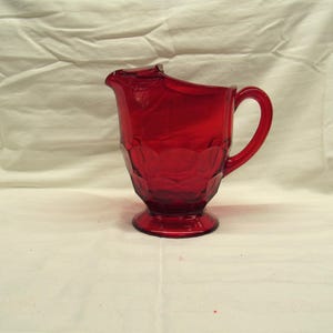 May include: A ruby red glass pitcher with a handle and a spout. The pitcher has a textured, geometric pattern on its body and sits on a round base. The handle is curved, and the pitcher is set against a white background.