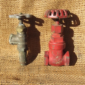 May include: Two vintage industrial valves. One is a tarnished silver color with a star-shaped handle. The other is a bright red with a round handle. Both valves are made of metal and appear to be well-used.