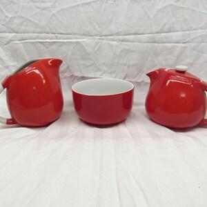 May include: A red ceramic tea set including a teapot with a lid, a creamer, and a bowl. The teapot and creamer have white handles. The bowl has a white interior. The set is on a white surface.