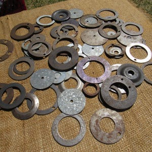 May include: A collection of various metal washers and circular components in shades of gray, silver, and brown. The assortment includes flat washers, some with holes, and others with raised centers, scattered on a textured brown surface.