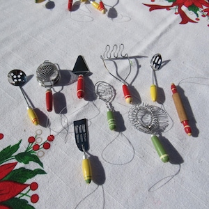 May include: A collection of miniature kitchen utensils, including a slotted spoon, whisk, and spatula. Each tool features a colorful handle with red, yellow, and green accents. The utensils are arranged on a white surface with floral patterns.