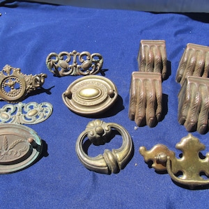 May include: A collection of assorted brass furniture hardware including four claw-shaped feet, several ornate drawer pulls with intricate scrollwork, and oval-shaped backplates featuring embossed wheat motifs and decorative border patterns.