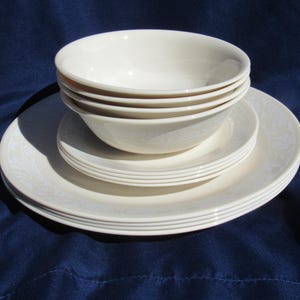May include: A stack of off-white ceramic bowls and plates. The bowls are stacked on top of a plate, with additional plates beneath. The dinnerware has a subtle floral pattern and is set against a dark blue background.