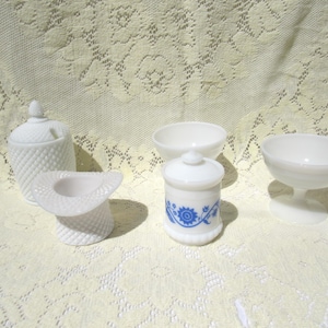 May include: A collection of white milk glass serving pieces, including a lidded sugar bowl, small bowls, a covered jar with blue floral design, and a stemmed dessert dish. The pieces are arranged on a lace tablecloth.
