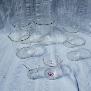 May include: A collection of clear glass beakers of various sizes, some with spouts and measurement markings. The beakers are arranged on a white, textured surface, showcasing their transparent design. Ideal for laboratory or scientific use.