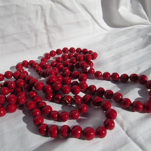 May include: A long, red beaded necklace with a glossy finish. The necklace is composed of numerous round beads, creating a bold and eye-catching accessory. The beads are approximately 0.5 inches in diameter. The necklace is displayed on a white, textured surface.
