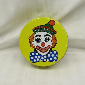 May include: A round, yellow tin featuring a cartoon clown face. The clown has orange curly hair, a red nose, and a smiling red mouth. It wears a green hat with a red pom-pom and a blue bow tie with white polka dots. The tin is likely a container.