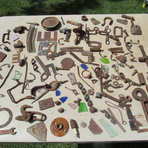 May include: A large collection of assorted rusty metal hardware, mechanical parts, and small fragments of colored glass scattered across a surface. The items include various hooks, gears, brackets, washers, a wrench, and small shards of blue and green glass.