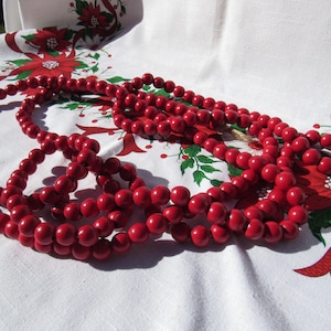 May include: Several strands of bright red wooden beads are draped across a white tablecloth. The tablecloth is decorated with a Christmas floral pattern featuring red poinsettias, green leaves, and red ribbons. The beads are smooth and round.