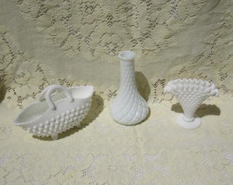 May include: Three pieces of white milk glass. A small basket with a handle, a vase with a textured diamond pattern, and a small pedestal dish with a ruffled edge. The objects are on a cream-colored lace tablecloth.