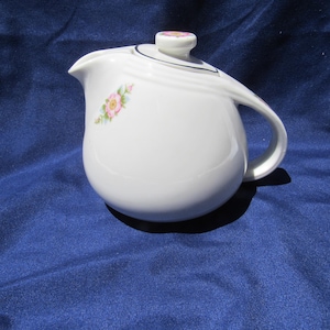 May include: A white ceramic teapot with a rounded body, a curved handle, and a matching lid. The teapot features a delicate floral pattern of small pink roses with green leaves on the side and a single matching rose design on the lid knob.