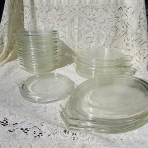 May include: A collection of clear glass plates and shallow dishes arranged in three separate stacks. Each piece features a smooth, transparent surface and a small, integrated handle on one side, designed for serving or heating food items.