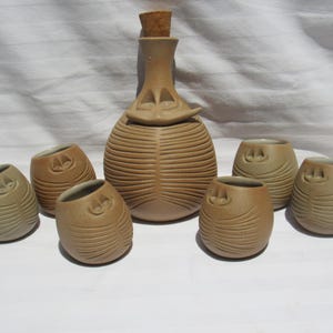 May include: A tan ceramic liquor set with a bottle and six small cups. The bottle has a cork stopper and a unique face-like design. The cups and bottle have a textured, ribbed design.