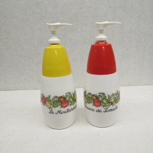 May include: Two white plastic condiment dispensers with pump tops. One has a yellow lid and the text Le Moutardier, the other has a red lid and the text Sauce de Tomate. Both feature a colorful botanical illustration of vegetables around the center.
