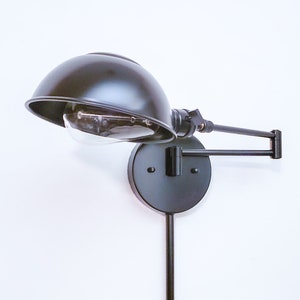 May include: Black wall-mounted lamp with a dome-shaped shade and adjustable arm. The lamp features a visible lightbulb and a round base. The cord extends downwards. The lamp is against a white background.