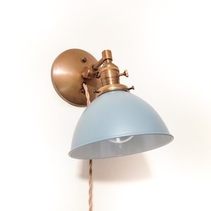 May include: A wall-mounted light fixture with a brass finish and a light blue metal shade. The fixture has a cord with a fabric-covered wire.