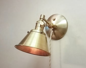 May include: A brass wall sconce with a cone-shaped shade and a white cord. The sconce is mounted on a white wall.