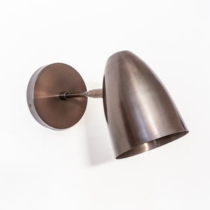 May include: A bronze wall-mounted light fixture with a round base and a long, narrow, angled shade.
