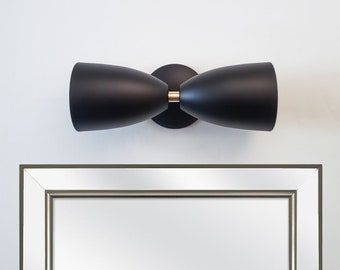 May include: A black wall sconce with two light fixtures shaped like rounded cones. The sconce has a gold accent in the center.