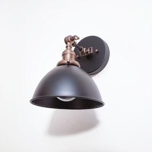 May include: A black metal wall sconce with a dome-shaped shade and a brass-colored arm. The sconce is mounted on a white wall.