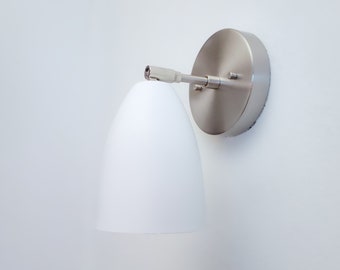 May include: A white wall-mounted light fixture with a white oval-shaped shade and a silver metal base.