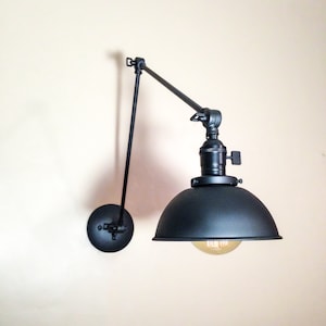 May include: Black metal wall-mounted light fixture with a swivel arm and a black metal shade. The light is on and the bulb is visible.