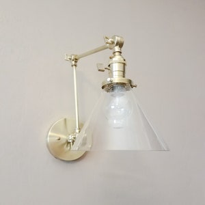 May include: A gold-toned wall sconce with a clear glass cone shade. The sconce has a swivel arm and a round base.