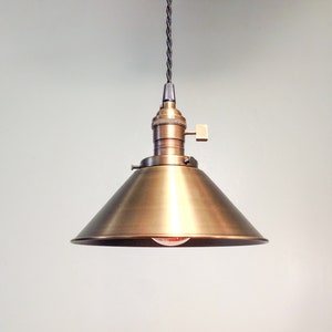 Puede incluir: Una l&aacute;mpara colgante de lat&oacute;n con una pantalla en forma de cono. La luz cuelga de un cable negro.