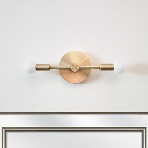 May include: A modern, brass-toned wall sconce with two white globe light bulbs. The fixture is mounted on a circular base and has a minimalist design. The sconce is mounted on a white wall, above a framed mirror.