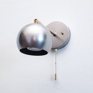 May include: A silver wall sconce with a round, globe-shaped shade and a pull chain switch. The sconce has a gold-colored arm and base.