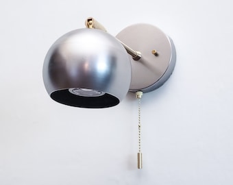 May include: A silver wall sconce with a round, globe-shaped shade and a pull chain switch. The sconce has a gold-colored arm and base.