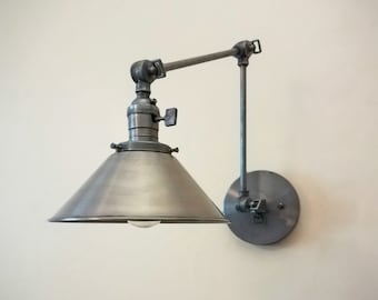 May include: A silver metal wall sconce with a cone-shaped shade and an adjustable arm. The sconce has a simple, industrial design.