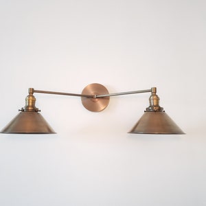May include: A bronze-toned, two-light wall sconce with a circular backplate and two adjustable arms. Each arm supports a conical shade. The fixture is mounted on a white wall, showcasing a vintage-inspired design.