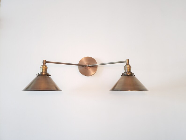 May include: A bronze-toned, two-light wall sconce with a circular backplate and two adjustable arms. Each arm supports a conical shade. The fixture is mounted on a white wall, showcasing a vintage-inspired design.