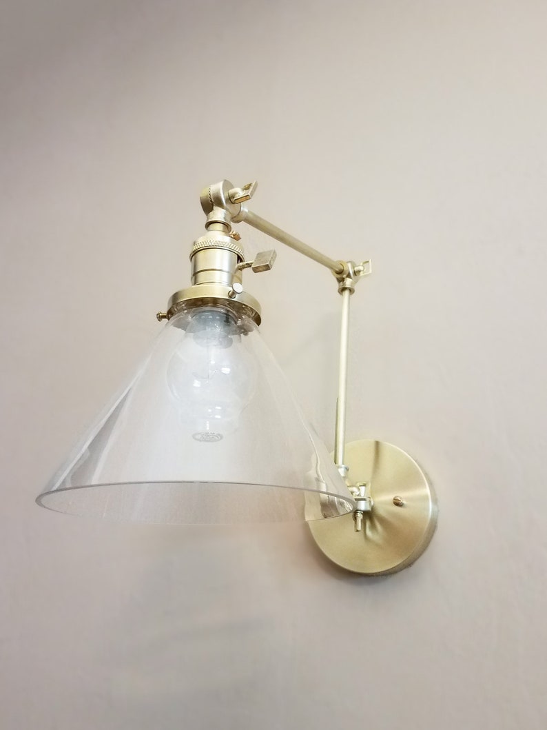 May include: A gold-toned wall-mounted light fixture with a clear glass shade. The shade is shaped like a cone and has a frosted finish. The fixture has a swivel arm that allows the light to be directed in different directions.