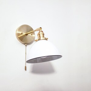 May include: A white and gold wall sconce with a white shade and a pull chain. The sconce has a round base and a curved arm.