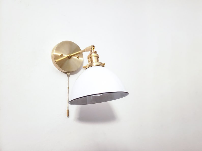 May include: A white and gold wall sconce with a white shade and a pull chain. The sconce has a round base and a curved arm.