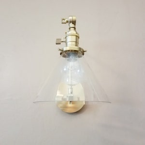 May include: A gold-toned wall sconce with a clear glass cone shade. The sconce has a simple design and is perfect for adding a touch of modern style to any room.
