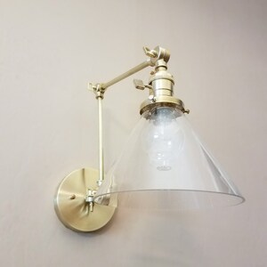 May include: A gold-toned wall sconce with a clear glass cone shade. The sconce has a swivel arm and a round base.