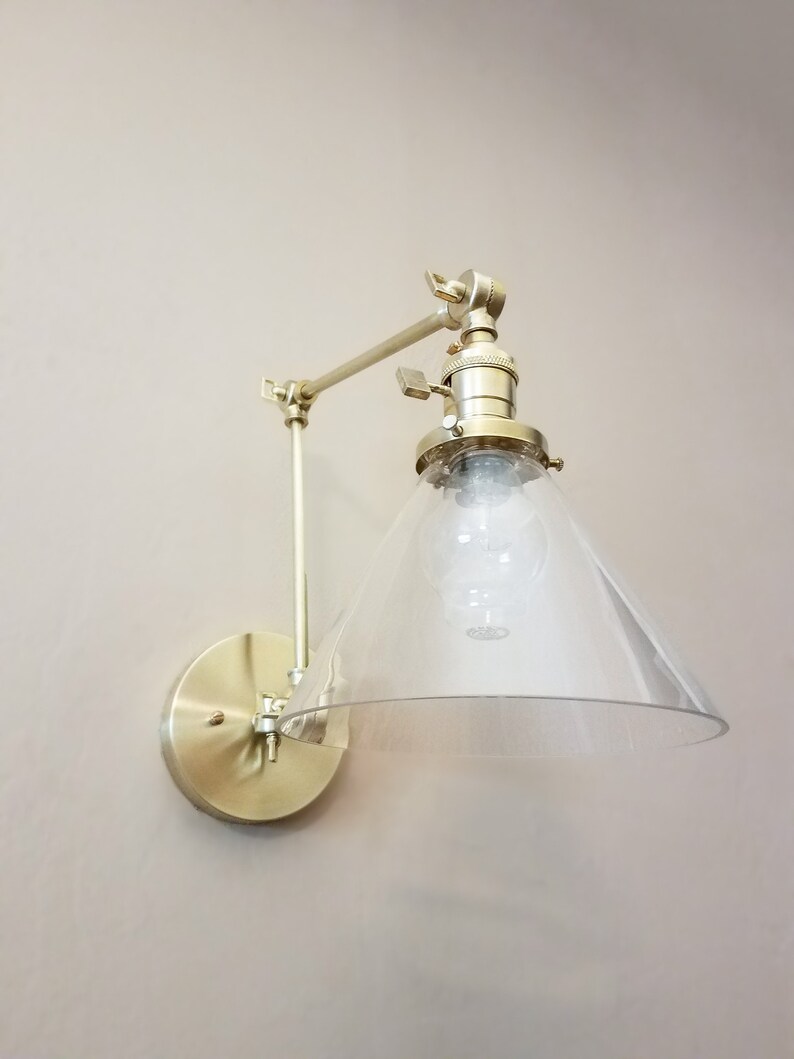 May include: A gold-toned wall sconce with a clear glass cone shade. The sconce has a swivel arm and a round base.