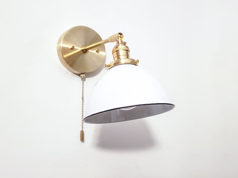 May include: A brass wall sconce with a white enamel shade and a pull chain switch. The shade has a black rim.