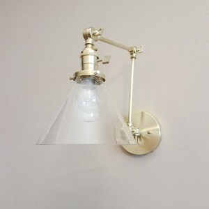 May include: A gold-toned wall sconce with a clear glass cone shade and an adjustable arm. The sconce has a simple, modern design and would be a great addition to any home.