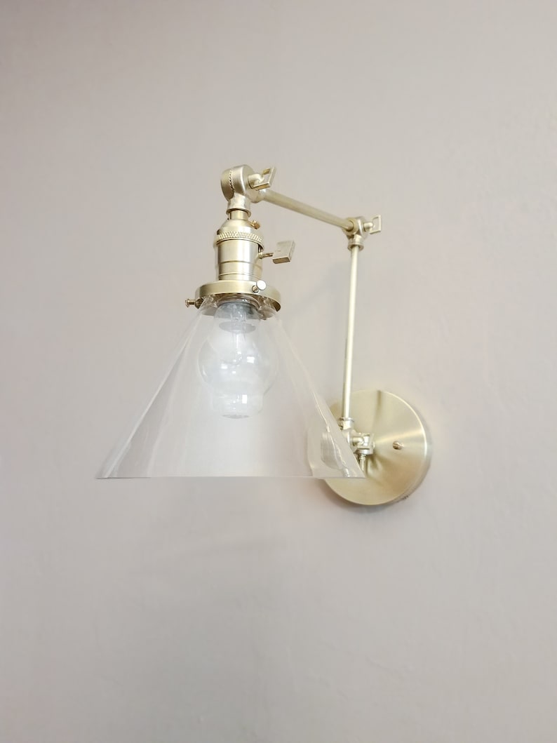 May include: A gold-toned wall sconce with a clear glass cone shade and an adjustable arm. The sconce has a simple, modern design and would be a great addition to any home.