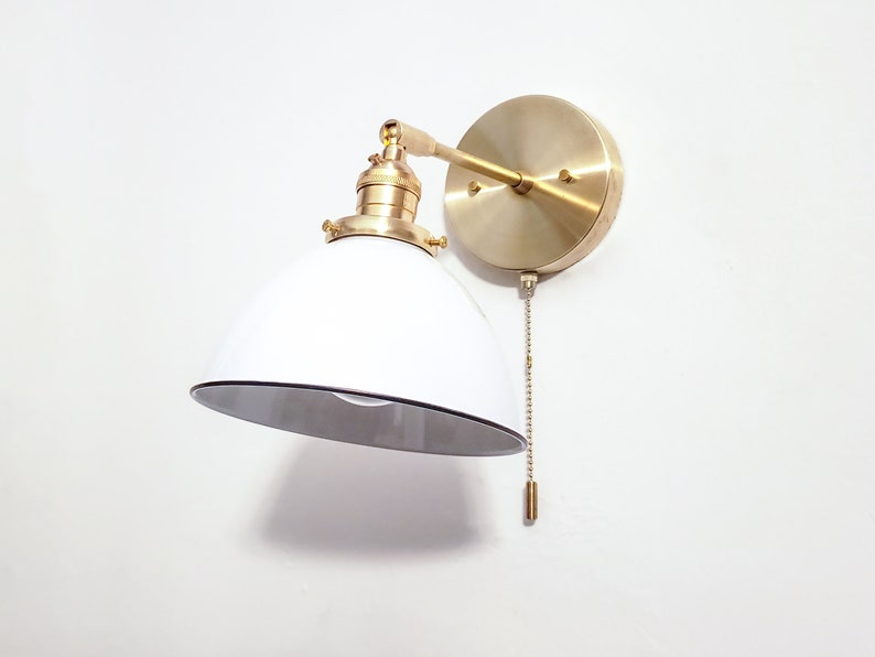 May include: A white and gold wall sconce with a pull chain switch. The sconce has a white, dome-shaped shade and a gold metal base.