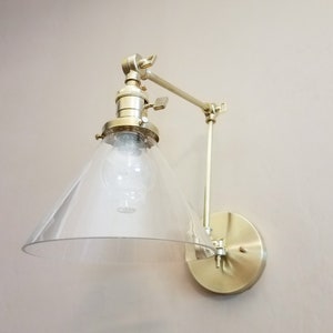 May include: A gold-toned wall-mounted light fixture with a clear glass shade. The shade is shaped like a cone and has a frosted finish. The fixture has a swivel arm that allows the light to be directed in different directions.