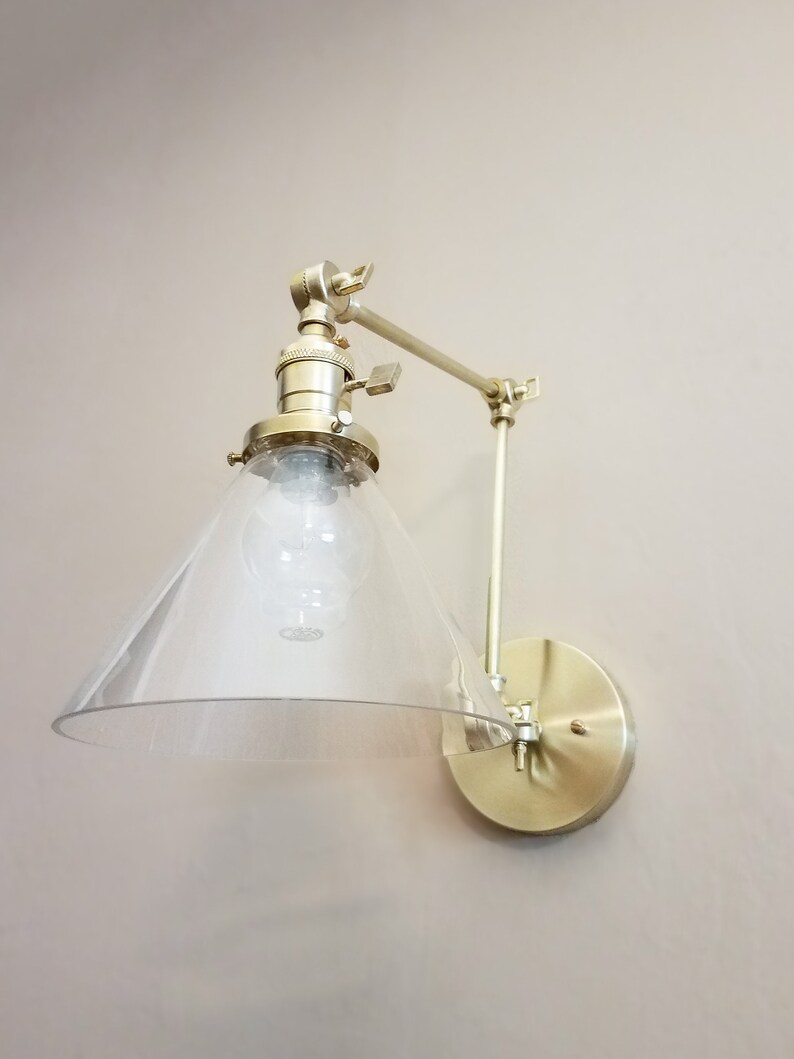 May include: A gold-toned wall-mounted light fixture with a clear glass shade. The shade is shaped like a cone and has a frosted finish. The fixture has a swivel arm that allows the light to be directed in different directions.