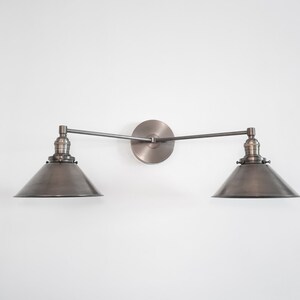 May include: A wall-mounted, two-light fixture with a vintage industrial design. The fixture features two conical shades and a circular backplate, all in a muted metallic finish. The arms are adjustable, and the fixture is mounted on a white wall.