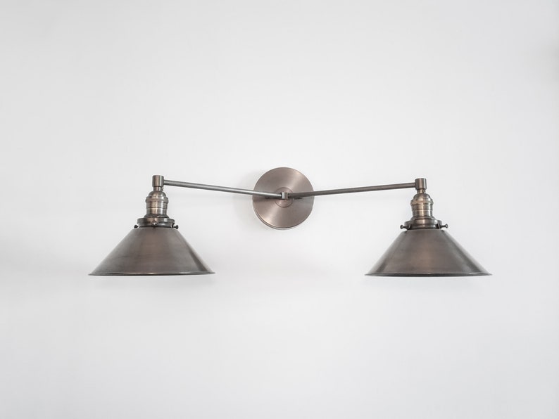 May include: A wall-mounted, two-light fixture with a vintage industrial design. The fixture features two conical shades and a circular backplate, all in a muted metallic finish. The arms are adjustable, and the fixture is mounted on a white wall.