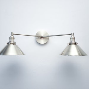 May include: A brushed nickel wall-mounted light fixture with two adjustable arms and conical shades. The fixture is mounted on a circular backplate. The shades are angled downward, and the fixture is against a white wall.