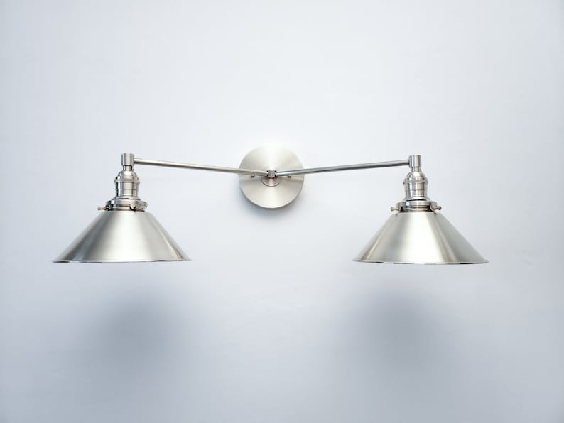 May include: A brushed nickel wall-mounted light fixture with two adjustable arms and conical shades. The fixture is mounted on a circular backplate. The shades are angled downward, and the fixture is against a white wall.